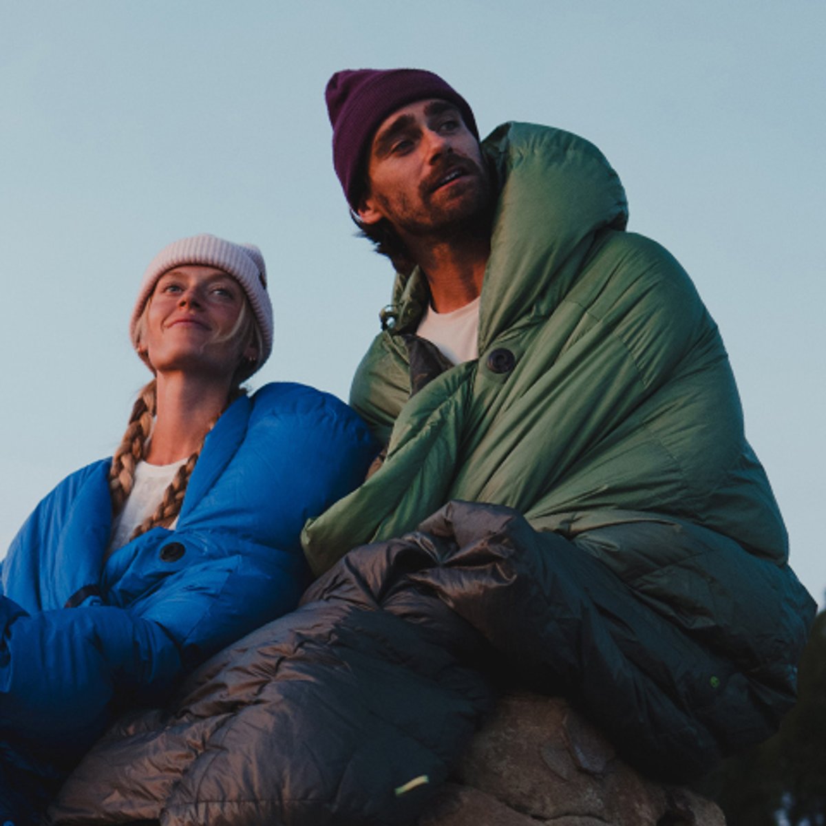 A man and a woman sit and watch the sunset, each wrapped in a cozy Marmot Wraptor sleeping bag. 