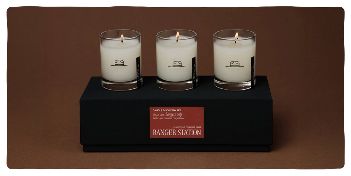 A rectangular box holding three lit candles in clear glasses with ranger hats printed on their exteriors. A sticker on the box says Candle Discovery Set: bangers only.