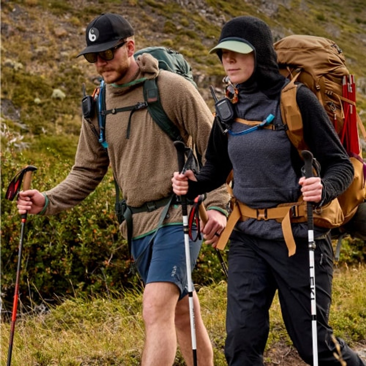 A grassy and rocky landscape with two hikers wearing backpacks walking on a trail. 
