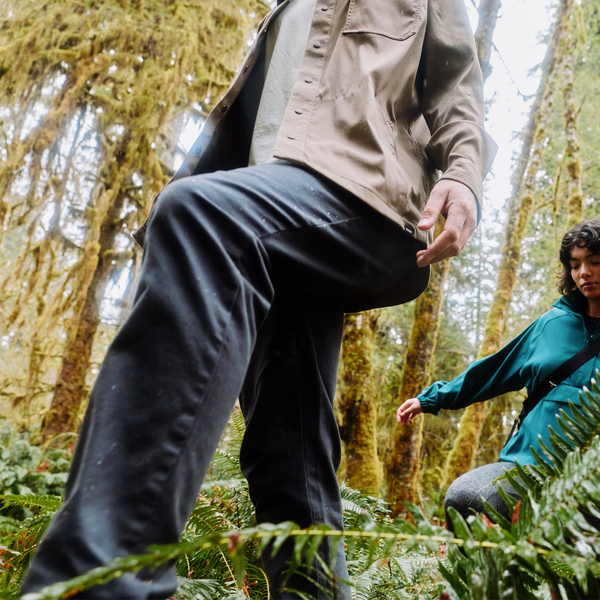 A person wears the prAna Stretch Zion pants, taking a large step in the forest, highlighting the 2-way stretch fabric.