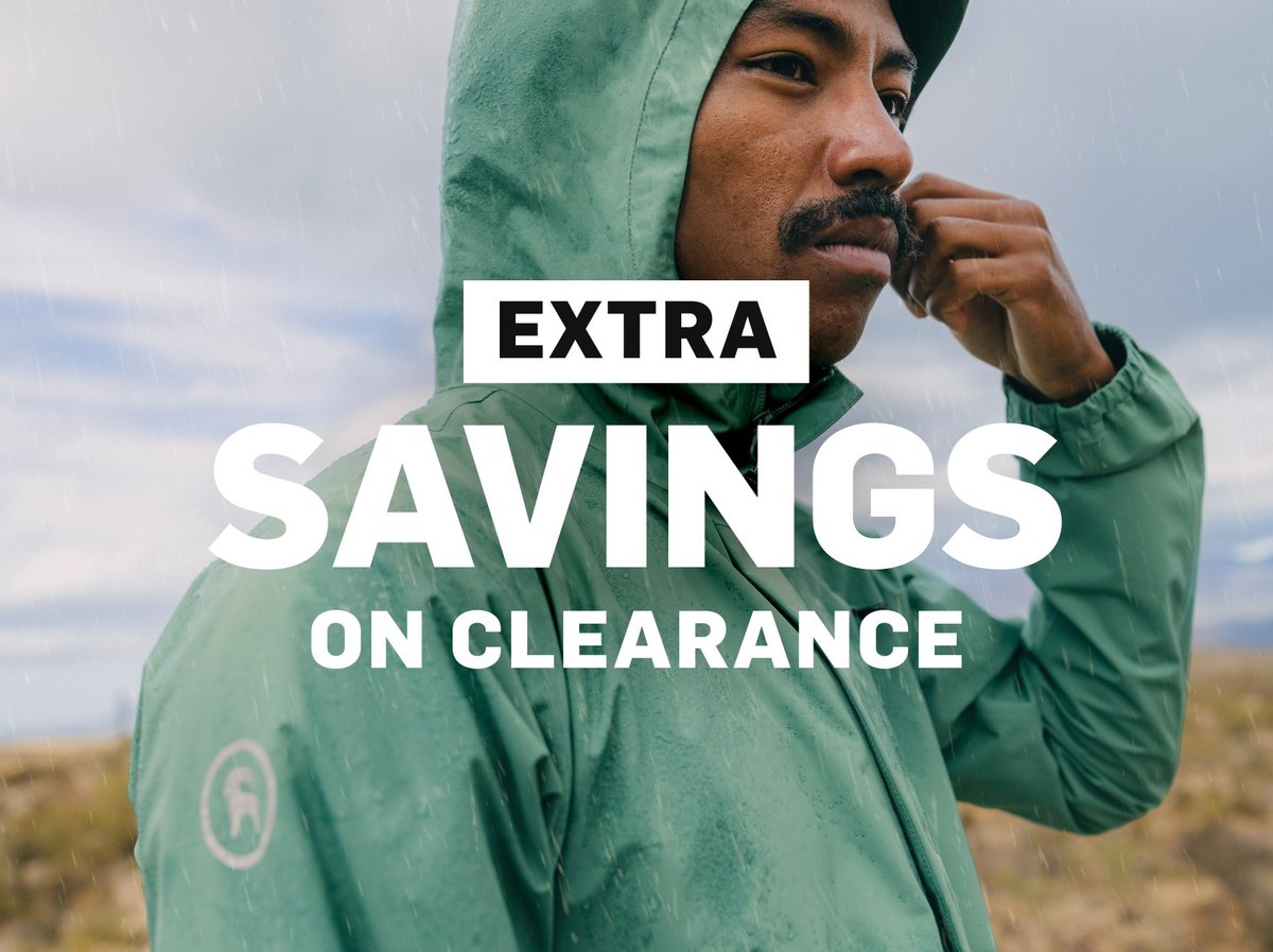 Hiker in a green BGA rain shell with the hood up, standing in light rain on open terrain: “Extra savings on clearance"