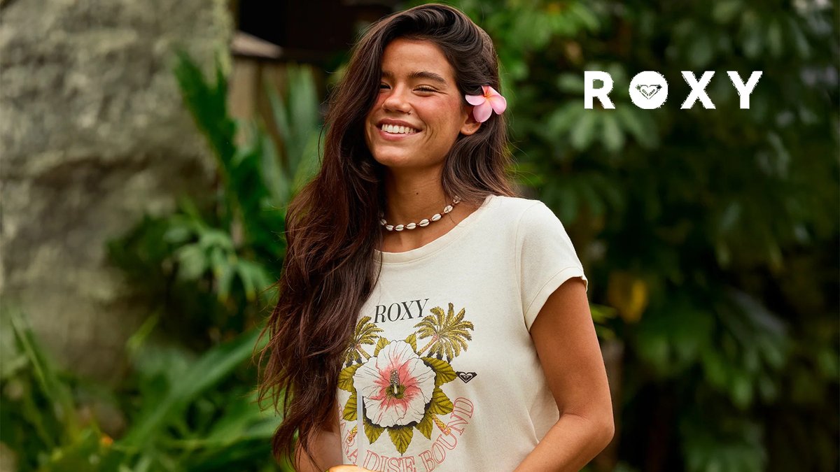 Smiling woman wearing a beige ROXY graphic t-shirt with palm tree and flower design, shell necklace, and pink hair clip in a tropical garden setting.