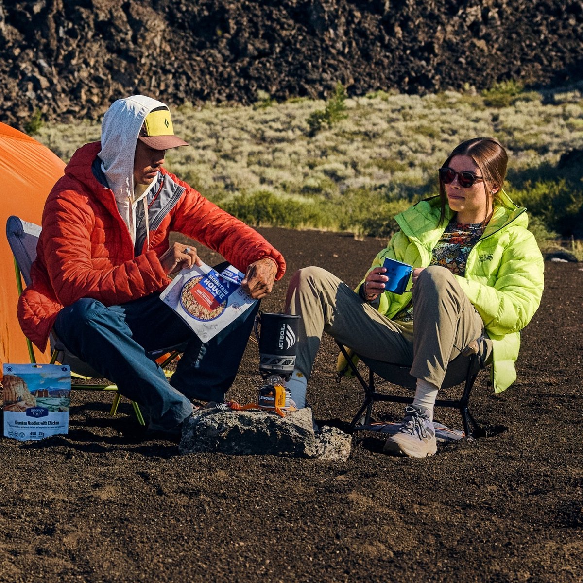 Two campers in folding chairs cooking a freeze-dried meal on a backpacking stove beside an orange tent and backpack on dark volcanic ground.