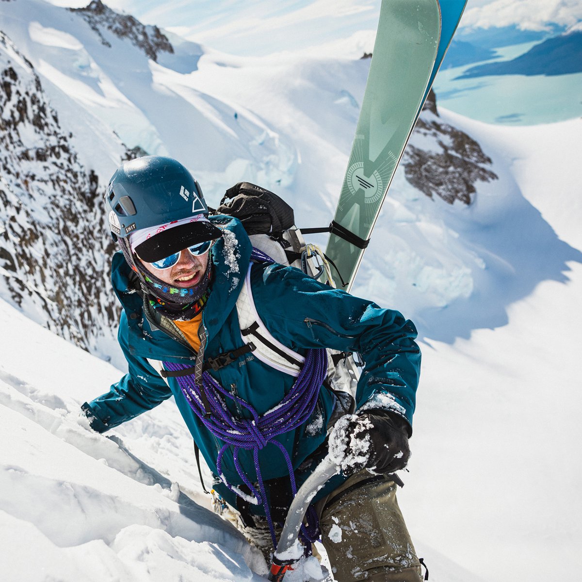 Man wearing an orange base layer under a teal jacket climbs a steep snow slope with skis and a rope strapped to his backpack, glacial terrain below.