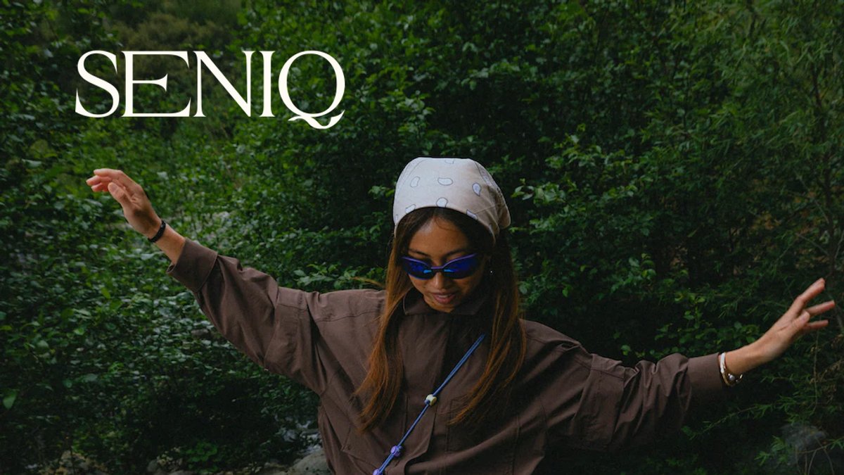 Woman wearing brown jacket, white cap, and blue sunglasses posing with arms spread against lush green foliage backdrop for Seniq.