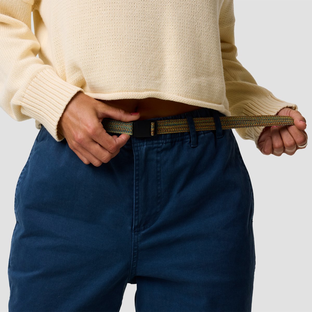 A woman adjusts the woven olive and tan striped canvas belt with a dark buckle on the BGA Venture Daily pant.