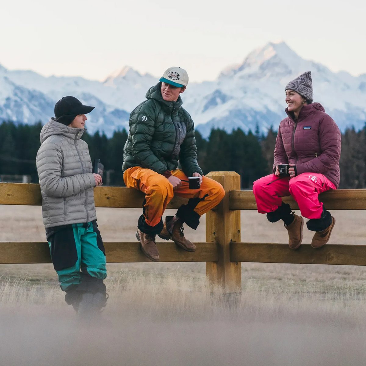 Three friends in insulated jackets and ski pants gather on a wooden fence for après with mugs, snow-covered peaks behind them.