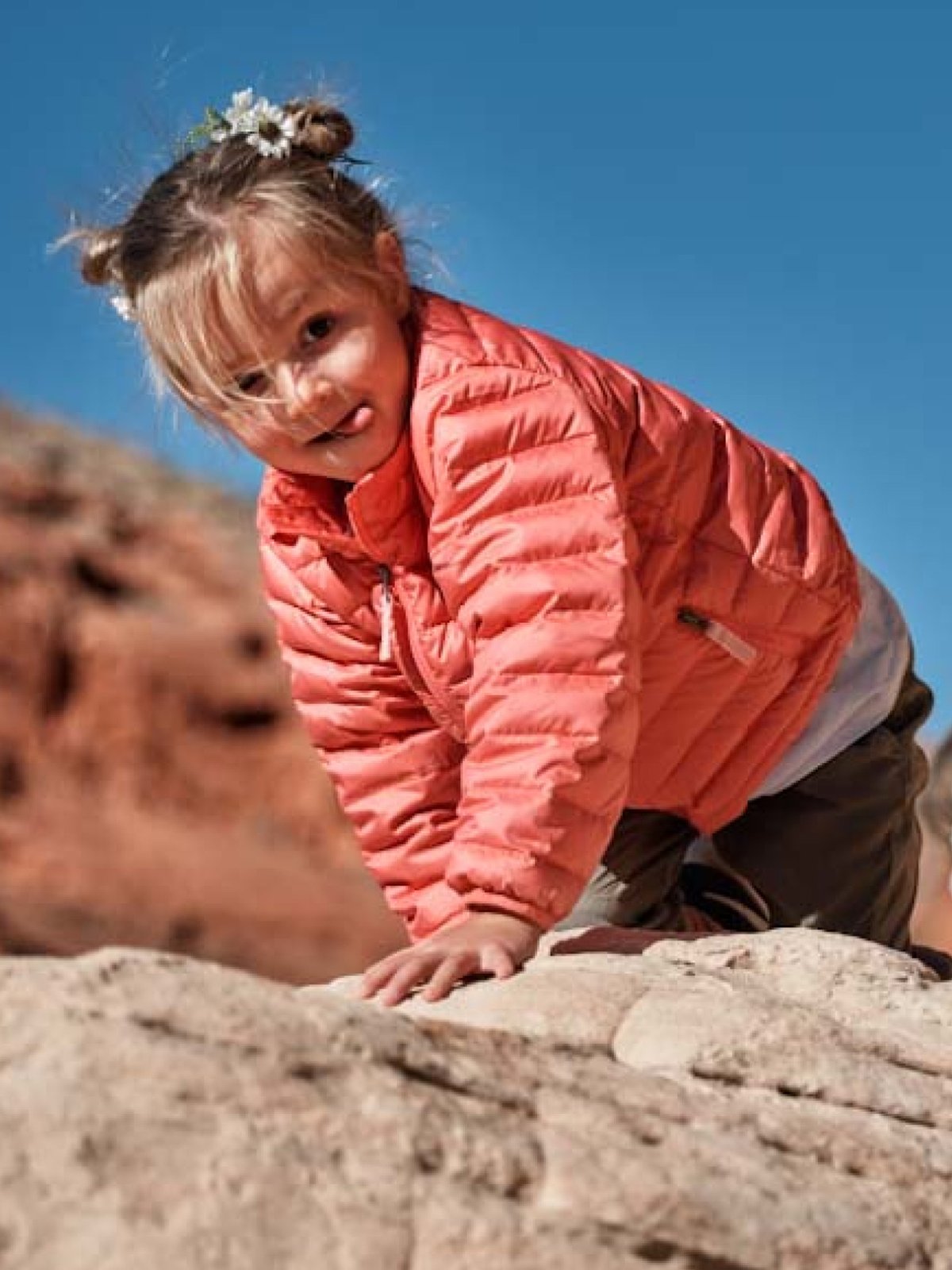A little girl in a bright coral puffy jacket climbs on a short granite boulder.