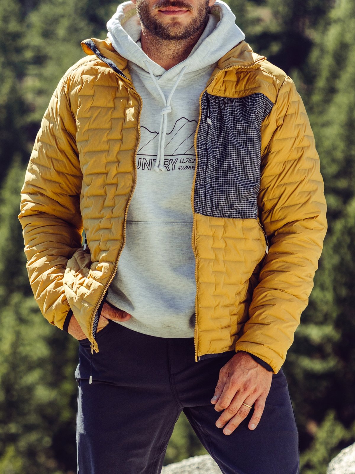 A person stands outdoors against a backdrop of trees, wearing a mustard yellow quilted jacket over a light gray hoodie with a graphic print and dark pants.