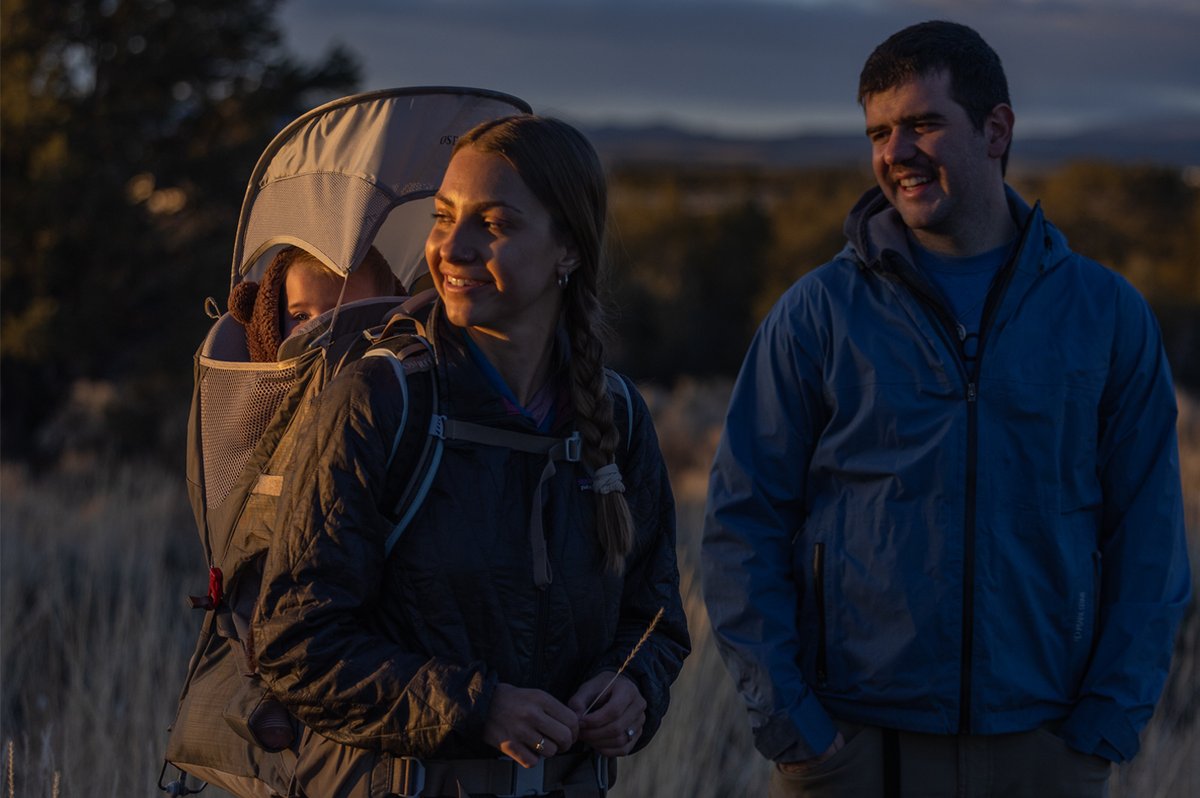 A couple with an Osprey carrier stands in a field at twilight.