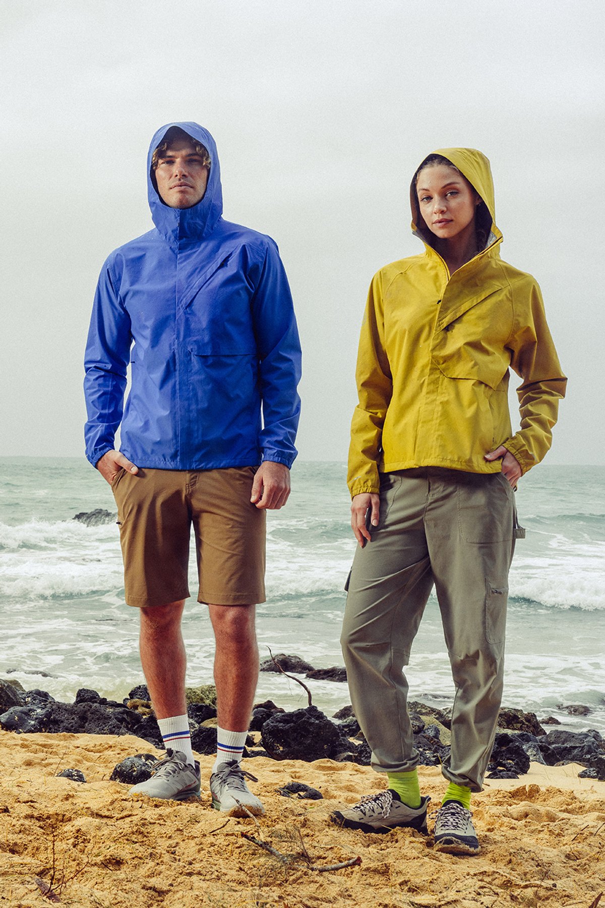 Man and woman wearing blue and yellow hooded rain jackets with hiking shorts and pants on rocky coastal beach