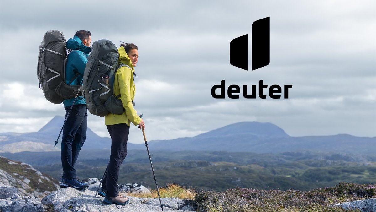 Two hikers wearing Deuter backpacks stand on a mountain summit overlooking a vast valley landscape on a cloudy day.