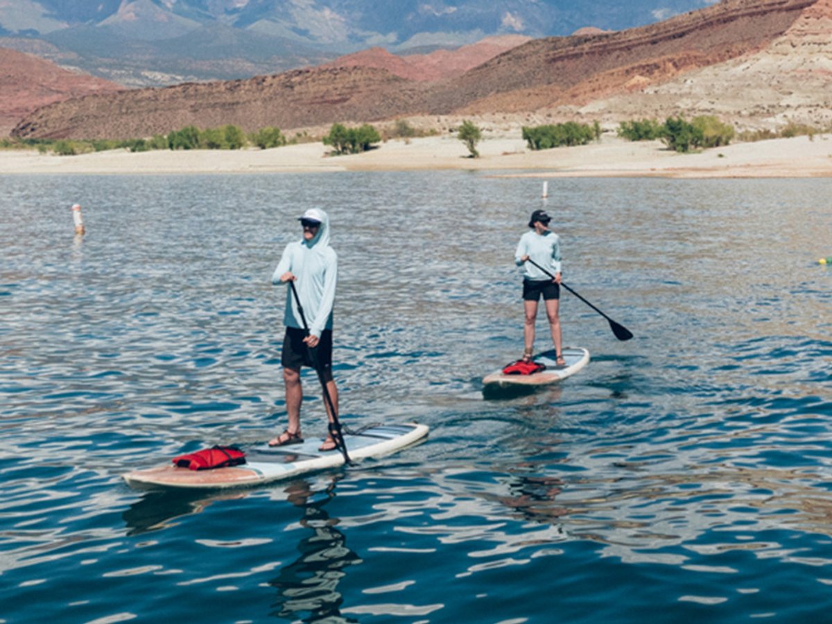 Two people in light blue sun hoodies and hats stand-up paddleboarding on a desert reservoir, with red sandstone cliffs and mountains behind them.