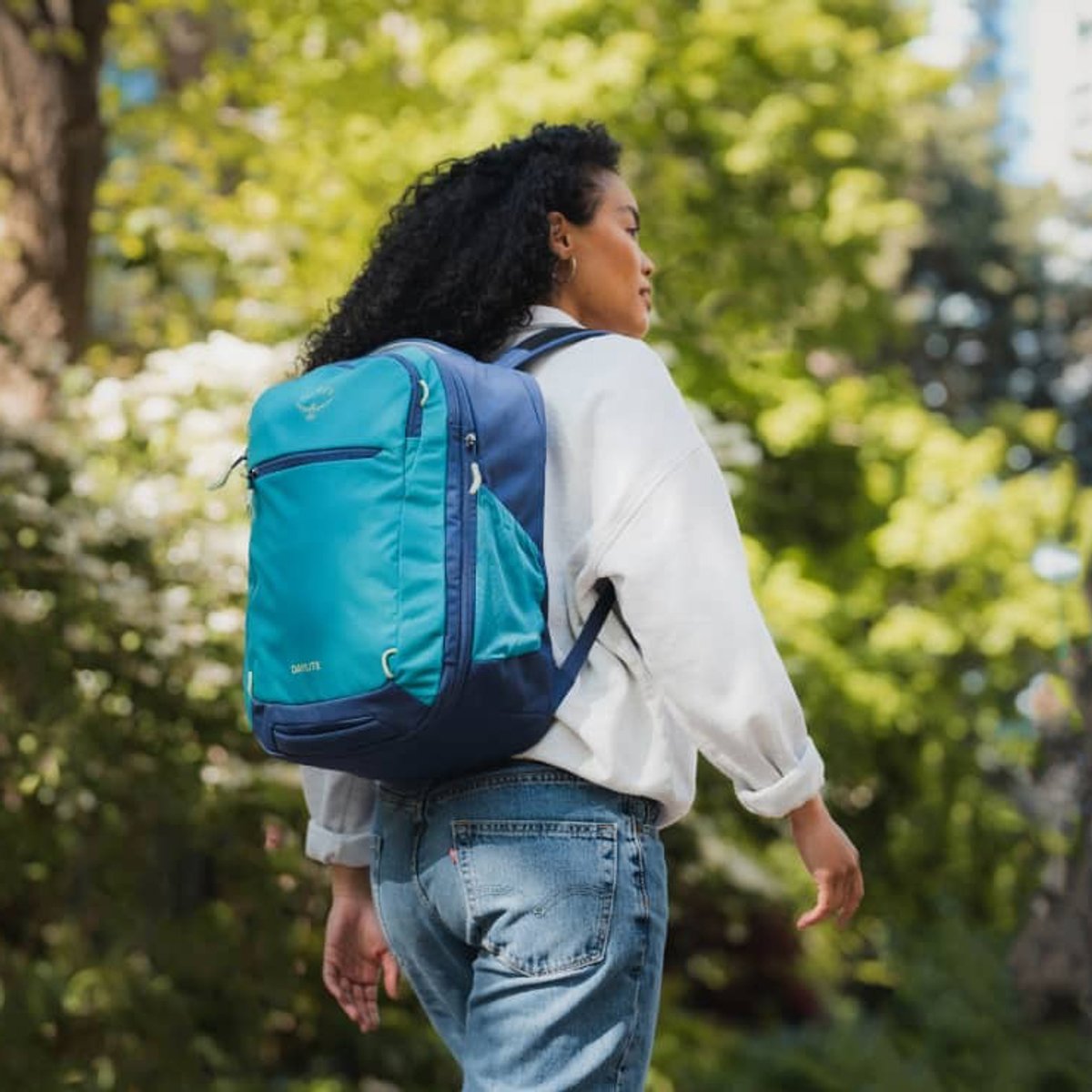 A woman wears an Osprey Travel backpack, walking down a shaded street path.