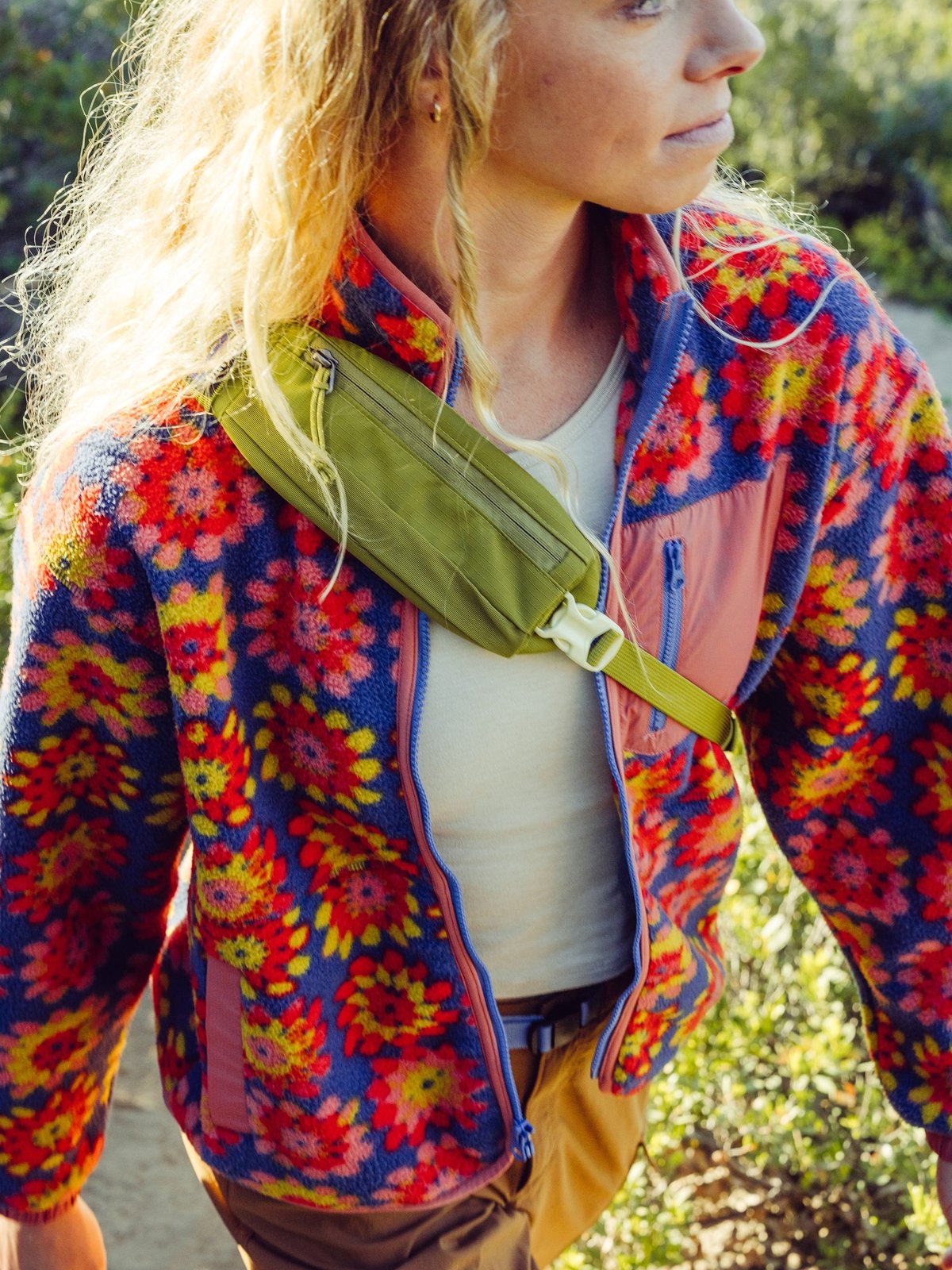 A person with long blonde hair hiking outdoors wearing a bright, colorful fleece jacket with a bold red, yellow, and orange floral pattern over a light shirt  and an olive green crossbody bag.