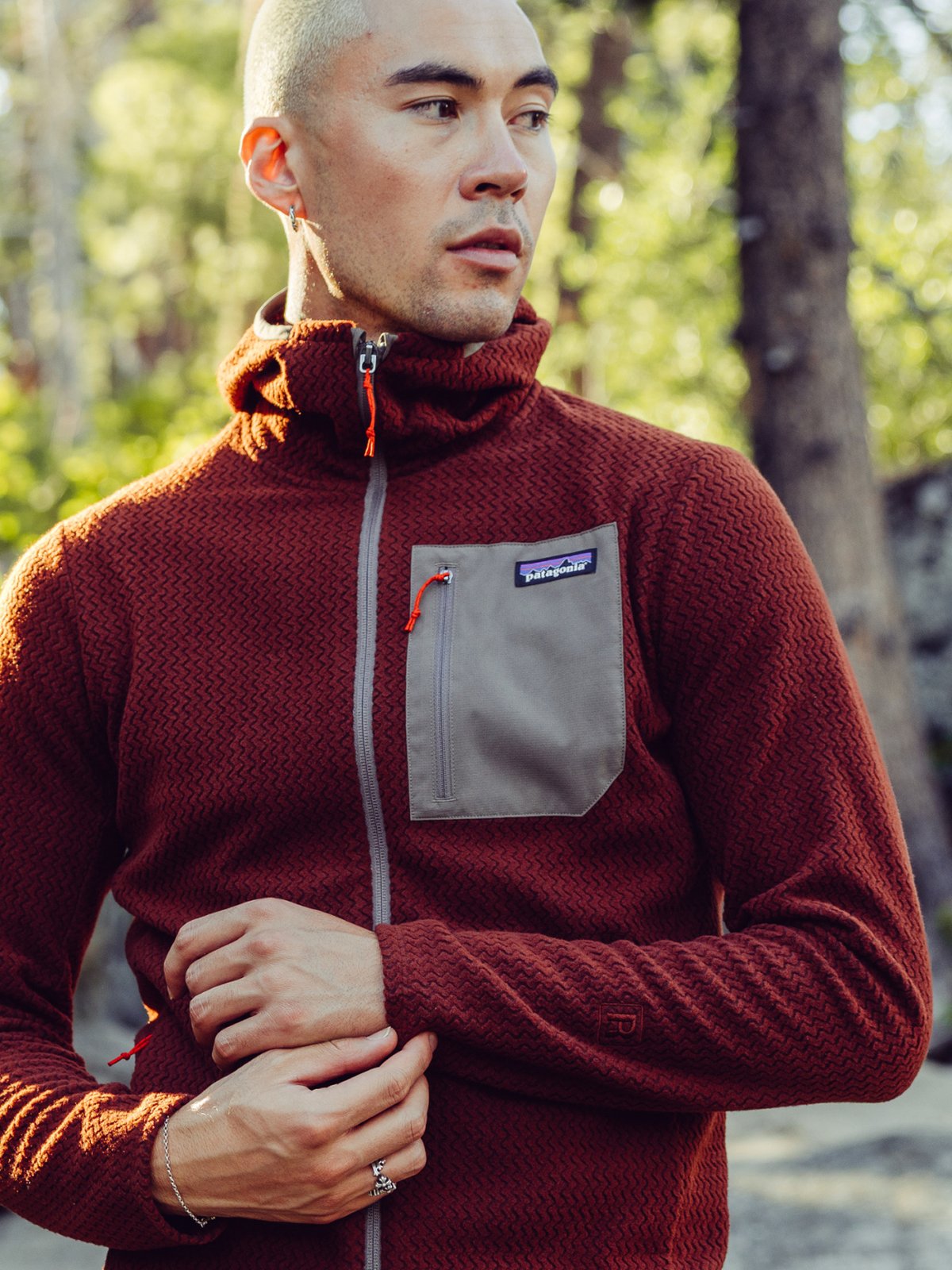 A hiker, wearing a red zip-up Patagonia fleece jacket with a light gray chest pocket, stands outdoors in a sunlit, wooded area.