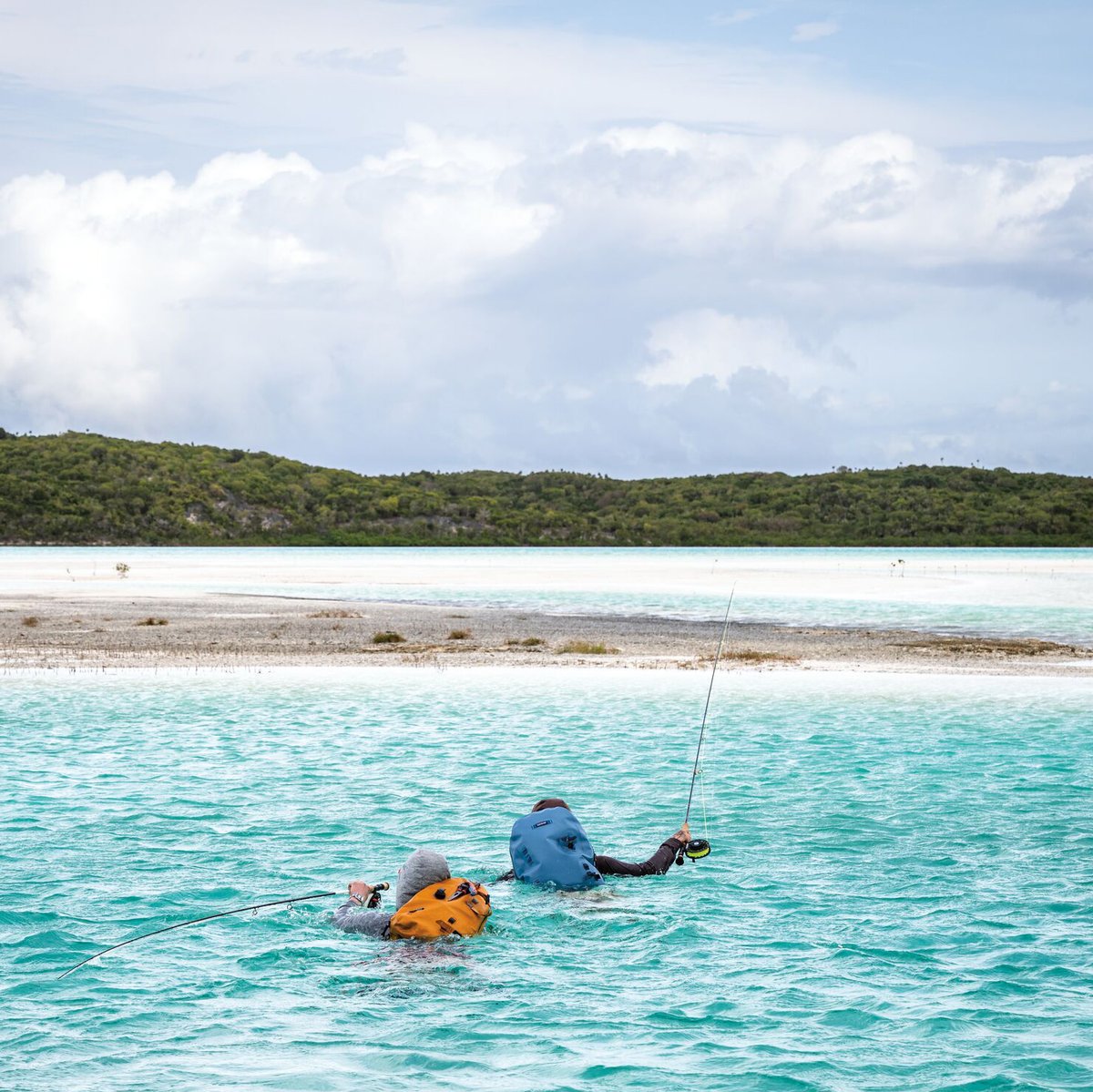 Two kayakers in yellow and blue kayaks paddle through turquoise tropical waters near a pristine white sand beach backed by lush green vegetation under a partly cloudy sky.