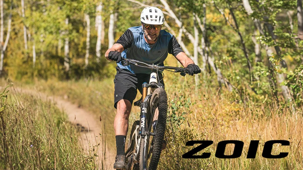 Male mountain biker riding a hardtail bike on a dirt trail through a forest wearing a white helmet and black athletic gear.