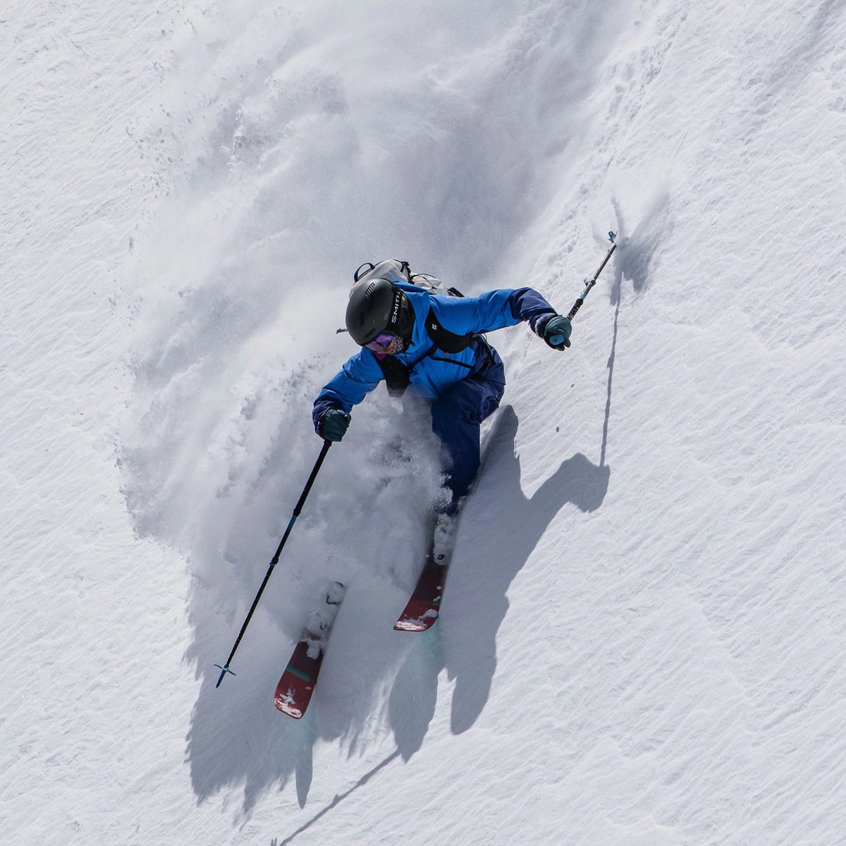 A person wearing a blue Black Diamond Recon jacket shreds down a slop, snow spraying in a super cool pattern behind them.