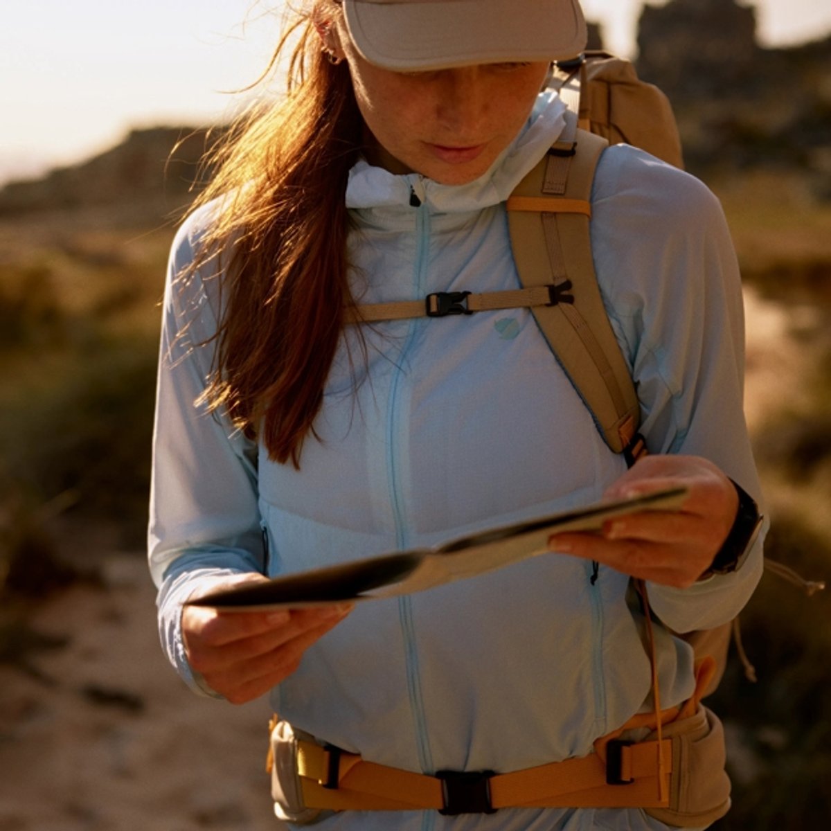 Woman wearing a light blue hiking jacket with tan backpack consulting a map while outdoors on a desert trail.