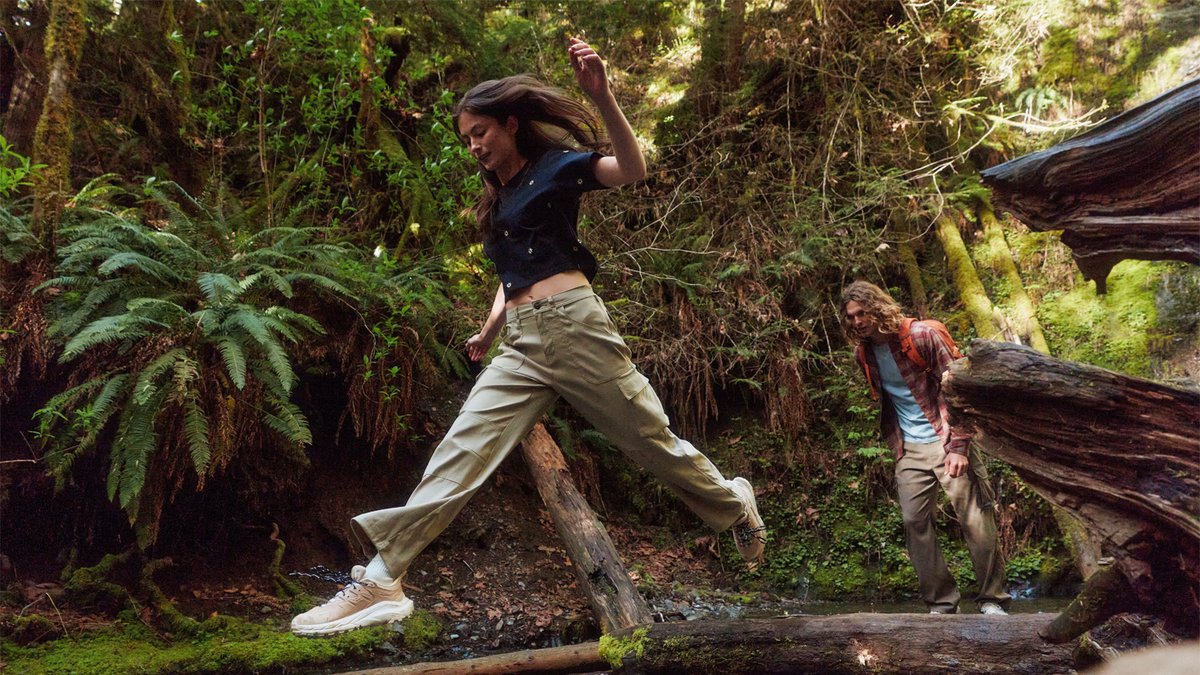 A man and woman jump over a log while wearing prAna Stretch Zion pants.