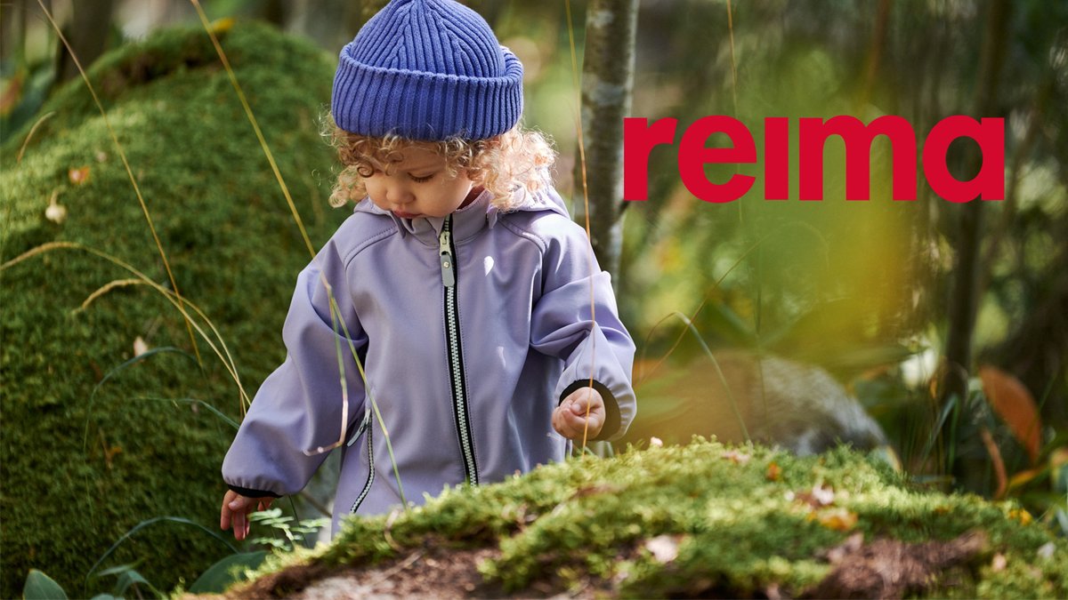 A toddler wearing a purple Reima rain jacket and blue knit beanie explores a mossy forest floor surrounded by green vegetation.