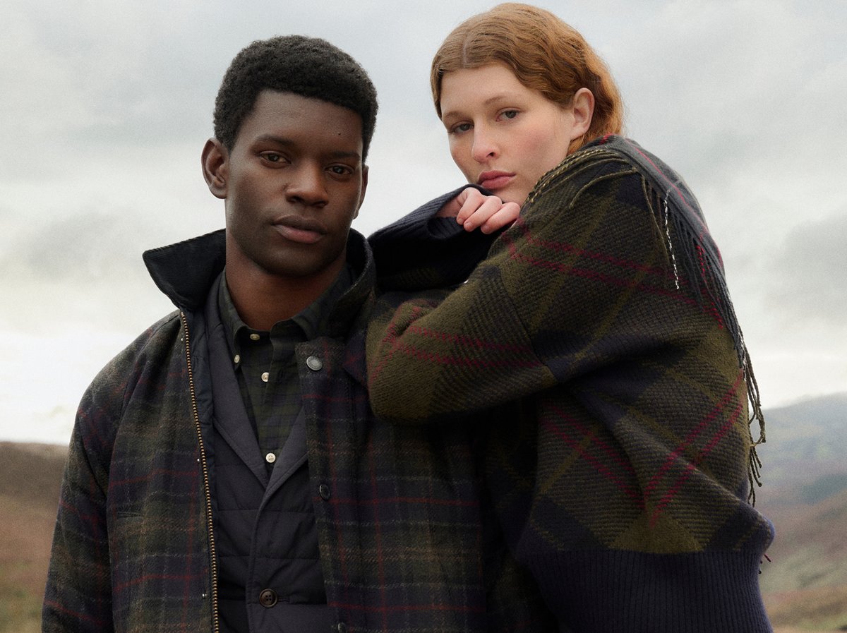 Two people wearing Barbour tartan outerwear and knitwear against a moody highland backdrop.