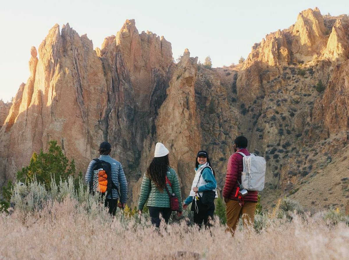 Four hikers with backpacks walk through a grassy field with large, sunlit rock formations in the background.