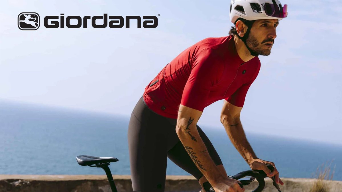 A male cyclist in red Giordana gear rides a road bike along a coastal cliff with blue ocean water in the background.