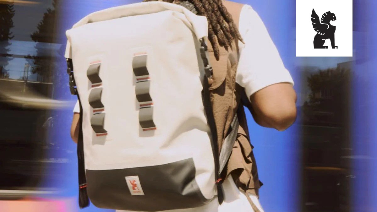 White and black Chrome waterproof backpack with ventilated straps and organization pockets worn by person with dreadlocks against blue background.