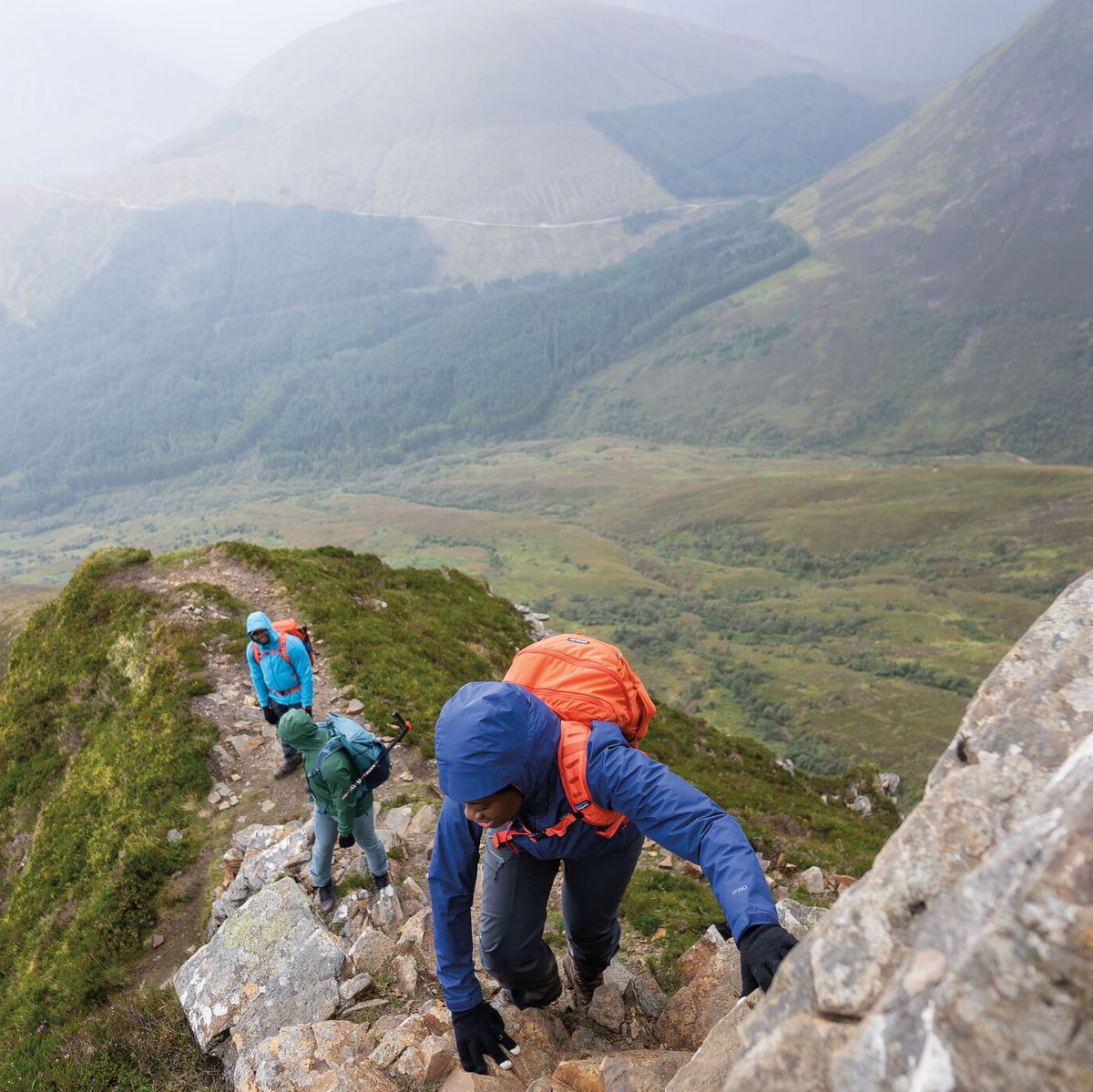 Three hikers in bright technical gear navigate a rocky mountain ridge with expansive valley views and forested slopes in the background during an outdoor adventure.