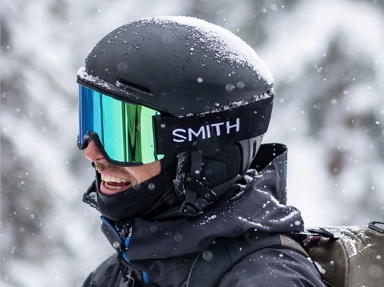   A man in snow gear smiles in the mountains. 