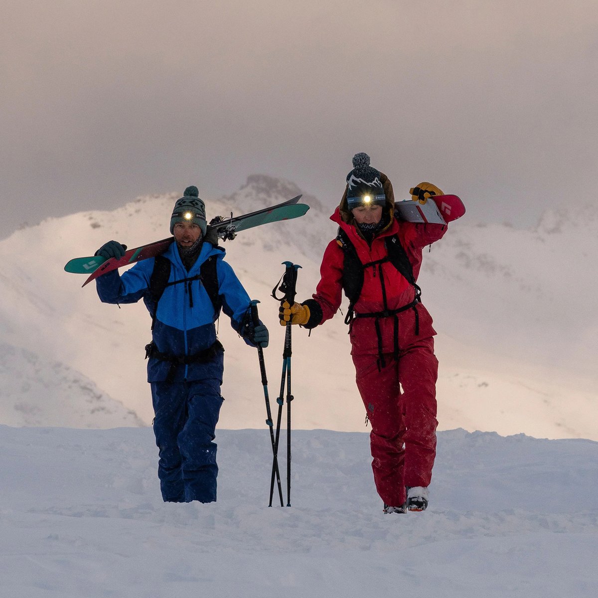 Two skiers walk through the snow, carrying skis over their shoulder.