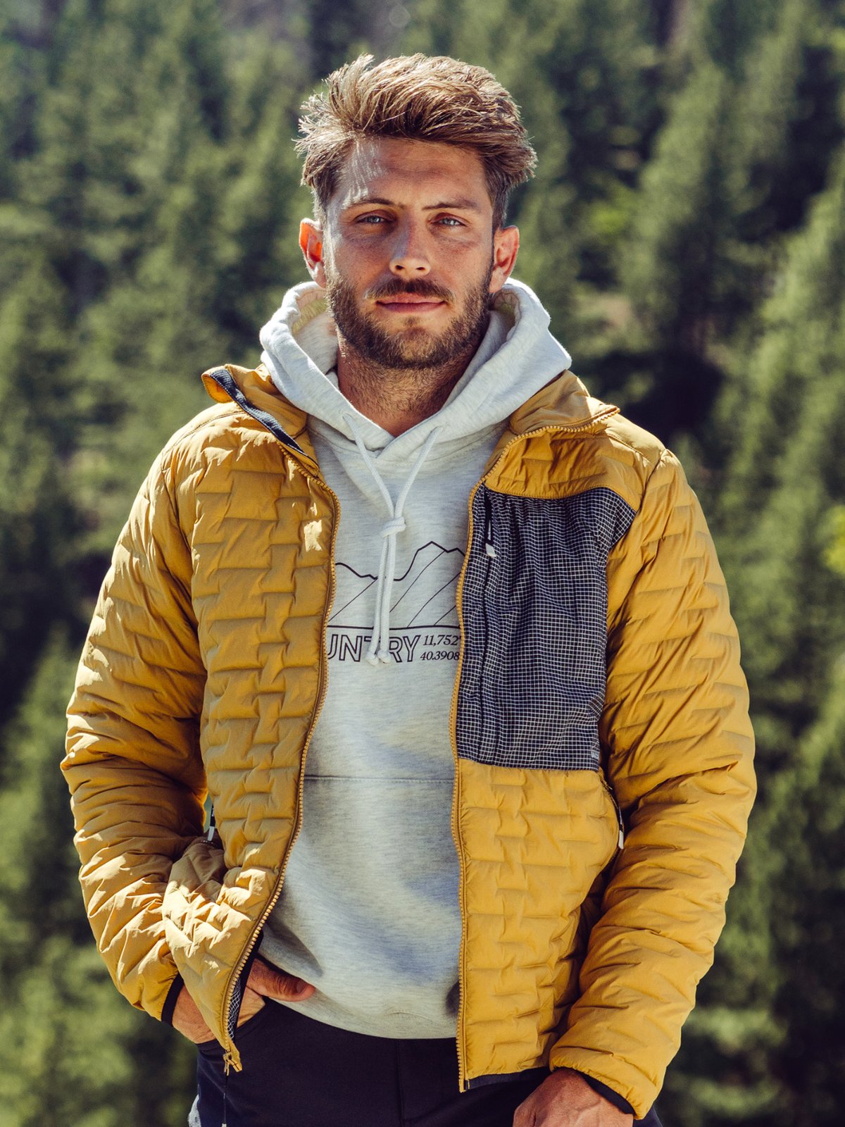 A man wearing a yellow quilted jacket over a light gray hoodie in a pine forest.