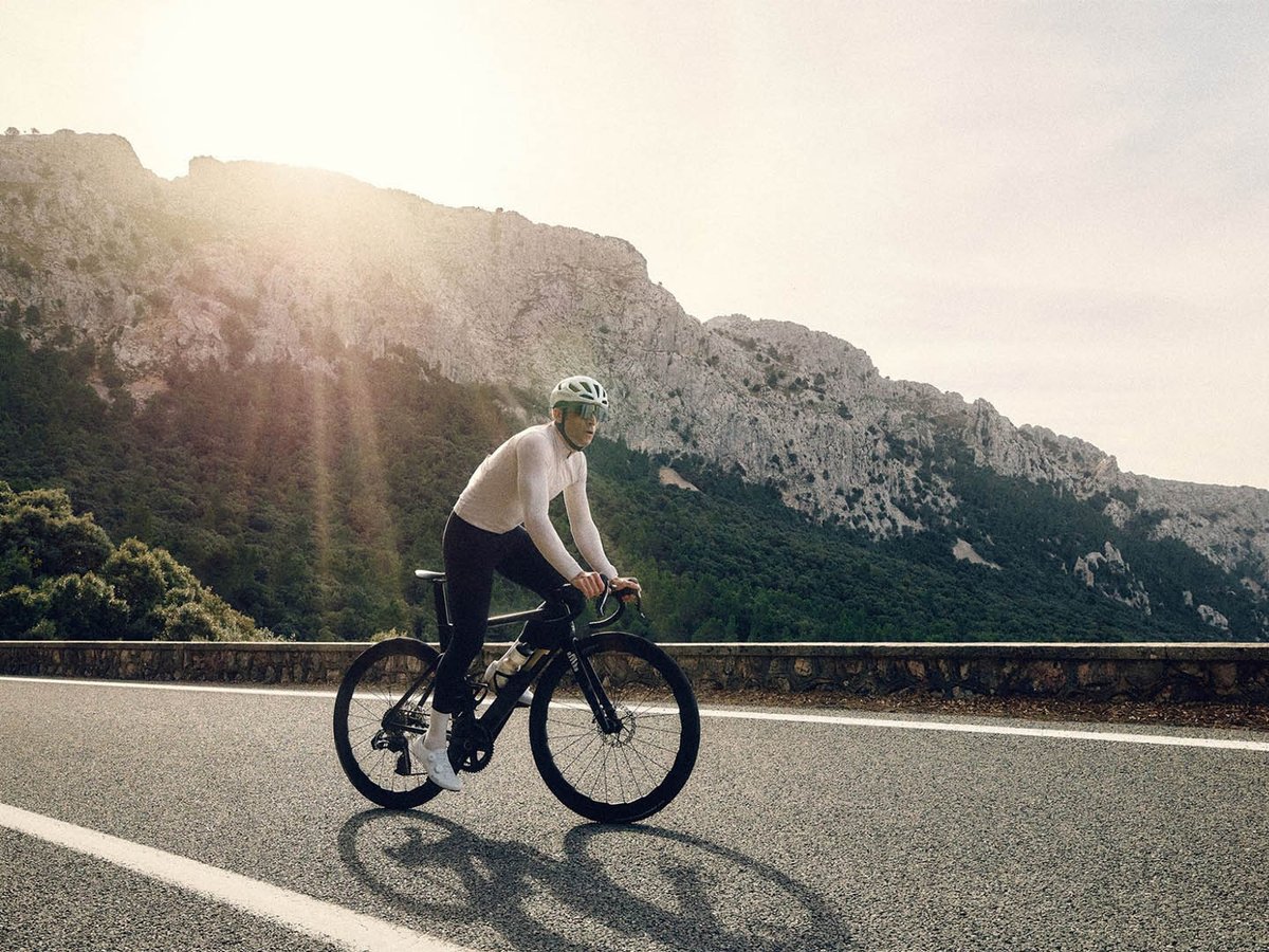 Male road cyclist in a white long-sleeve jersey and black bibs riding a black road bike on a mountain road, with a sunlit limestone cliff face behind him.