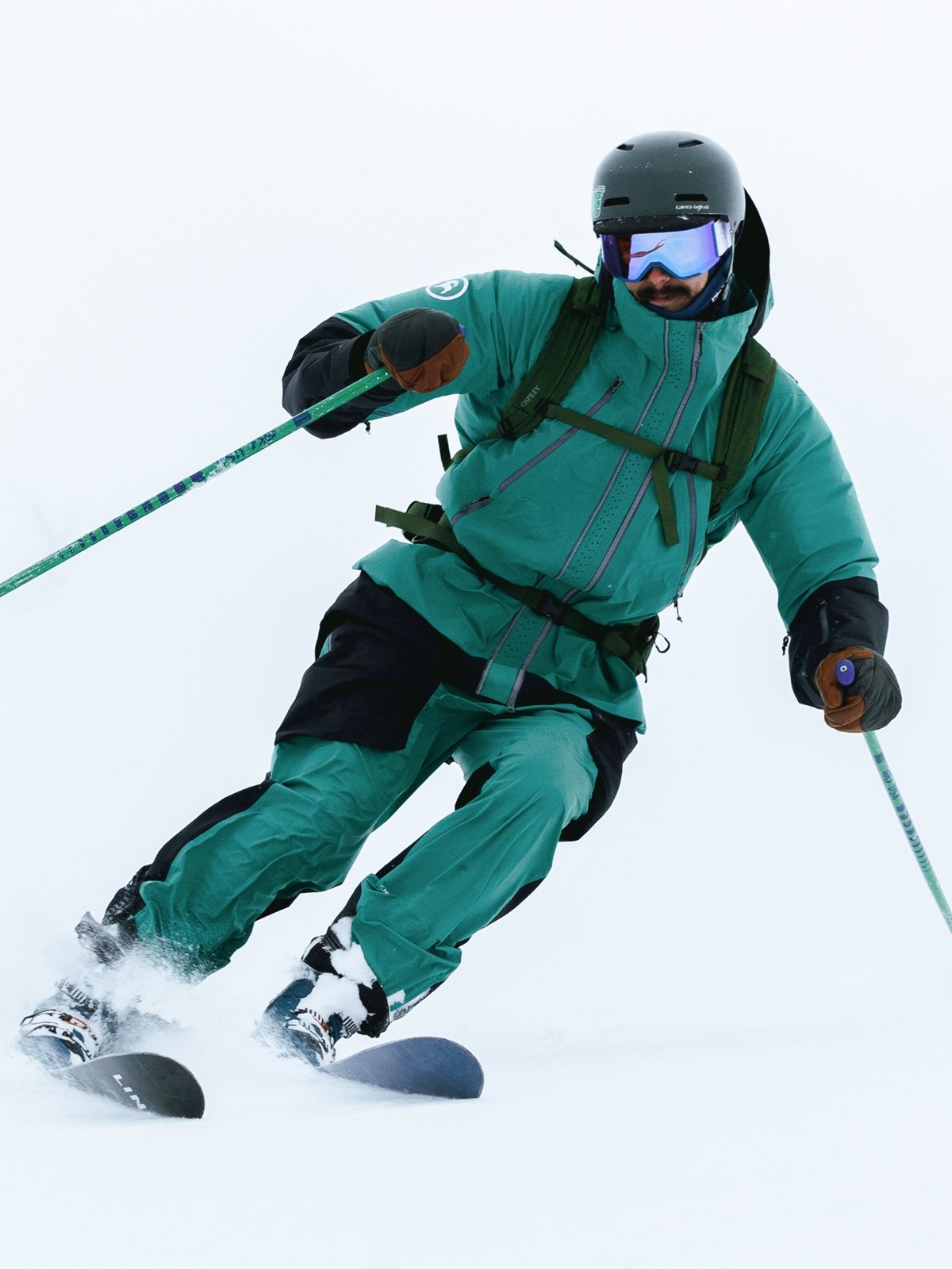 A ski tourer with a mustache wearing a green Backcountry Cardiac kit and backpack skins through falling snow.