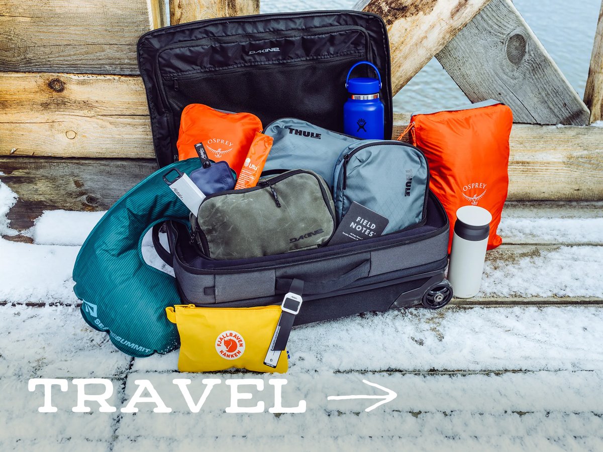 Open wheeled carry-on packed with packing cubes, a neck pillow, and a water bottle on a snowy wooden dock: “Travel.”