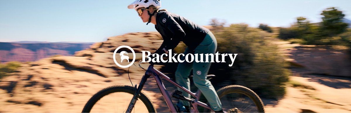 Woman riding a full-suspension mountain bike through a desert trail in Backcountry MTB apparel. Featuring the Backcountry logo.