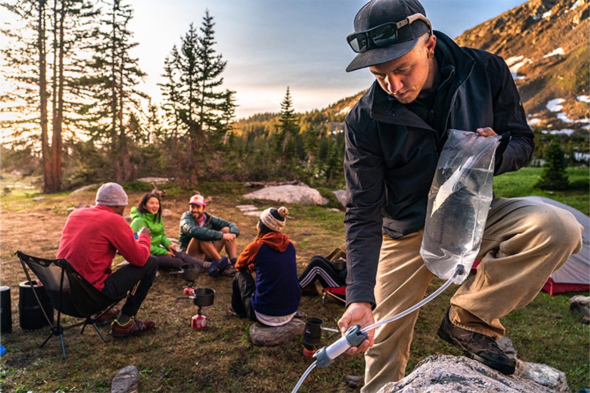 Man uses Platypus GravityWorks 4.0L filter system to fill containers at a campsite while a group relaxes in the background.
