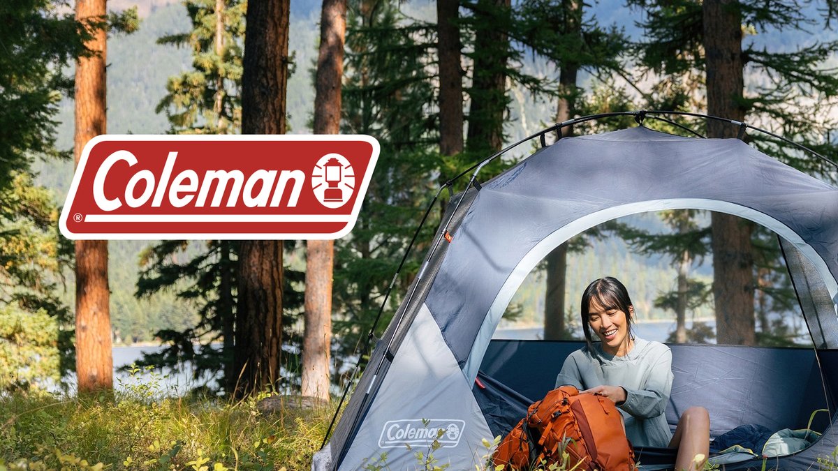 A woman sits inside a blue Coleman dome tent with an orange backpack, set up in a forested campsite near a lake with tall pine trees in the background.