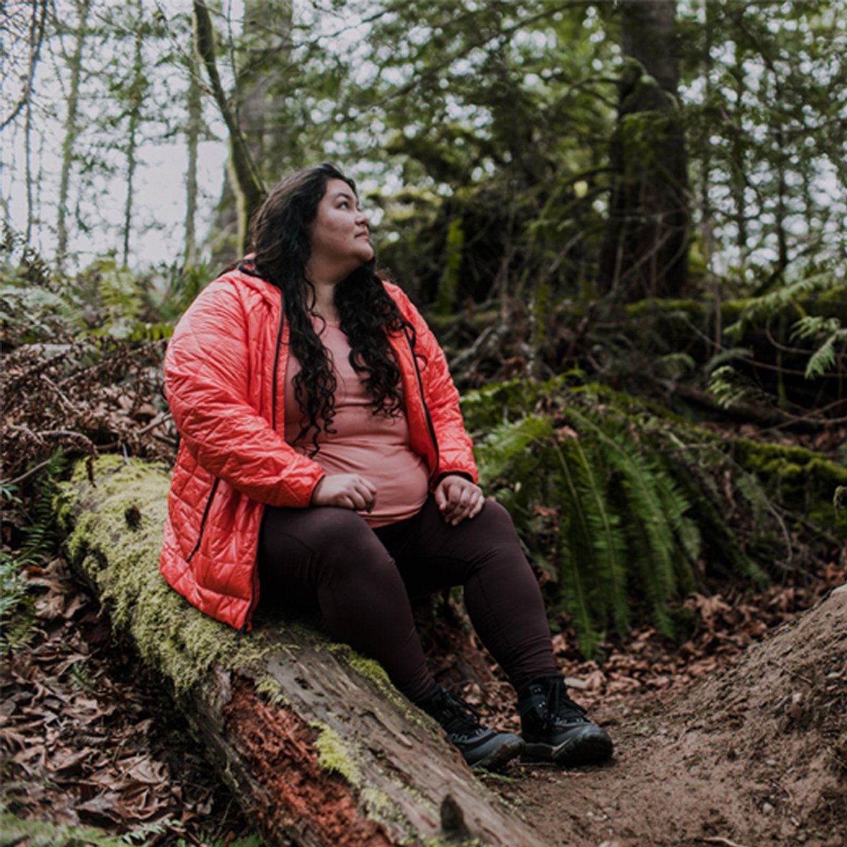 A woman wearing a Helium Insulated Hoodie sits on a fallen tree in a green forest. 