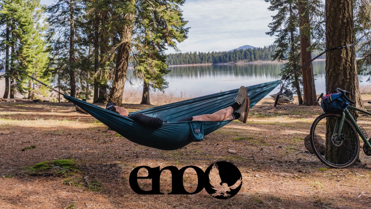 Person relaxing in a dark teal ENO hammock strung between trees at a lakeside campsite with a bicycle and mountain forest backdrop.
