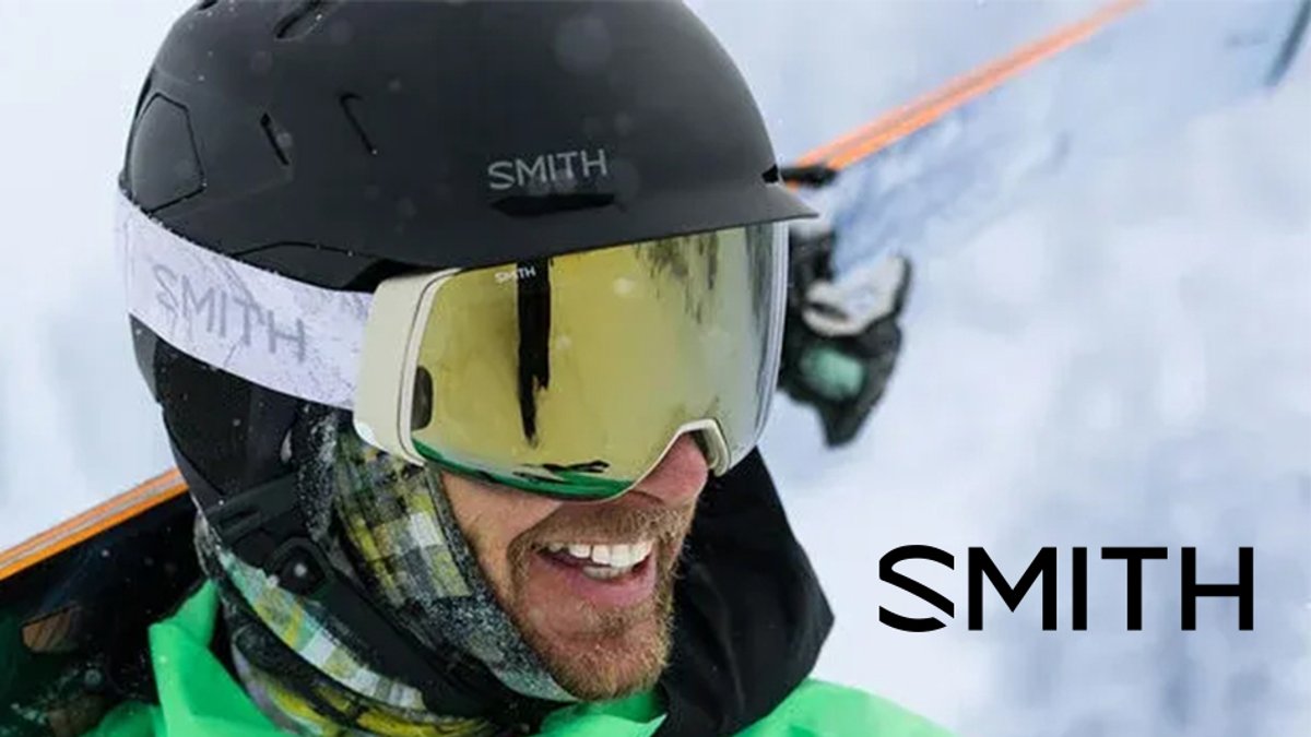 A smiling man wearing a black Smith helmet and gold-tinted goggles with a white strap stands against a snowy background.