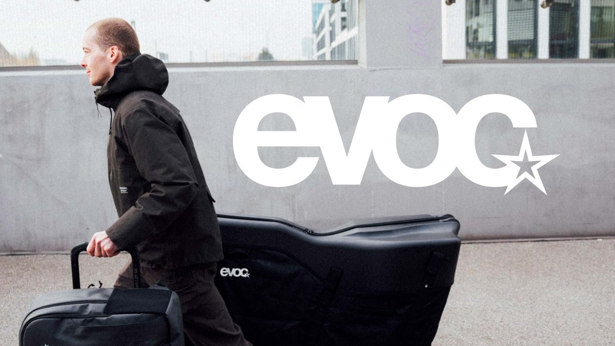 Man wearing black EVOC jacket walking with black EVOC bike travel bag and luggage in urban environment with modern buildings.
