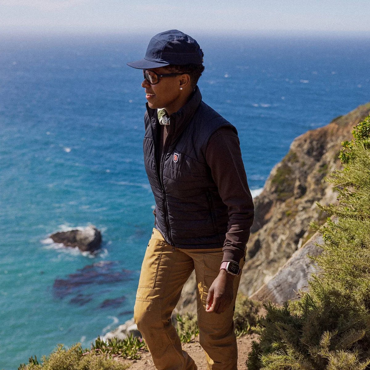 A woman walks on a coastal cliffside, wearing a Fjallraven Expedition Vest.