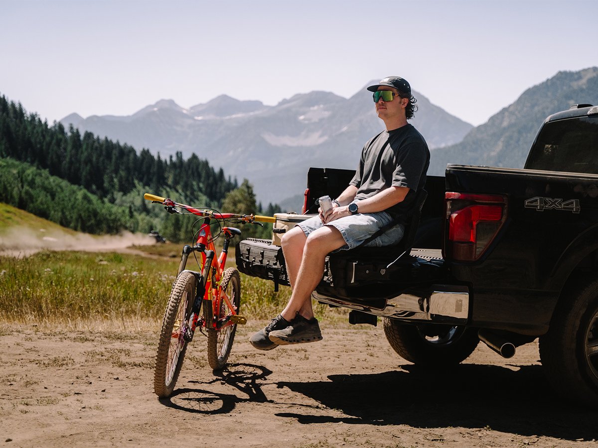 Person sitting on Cache Basecamp tailgate system with bike leaning nearby and mountains in the background.