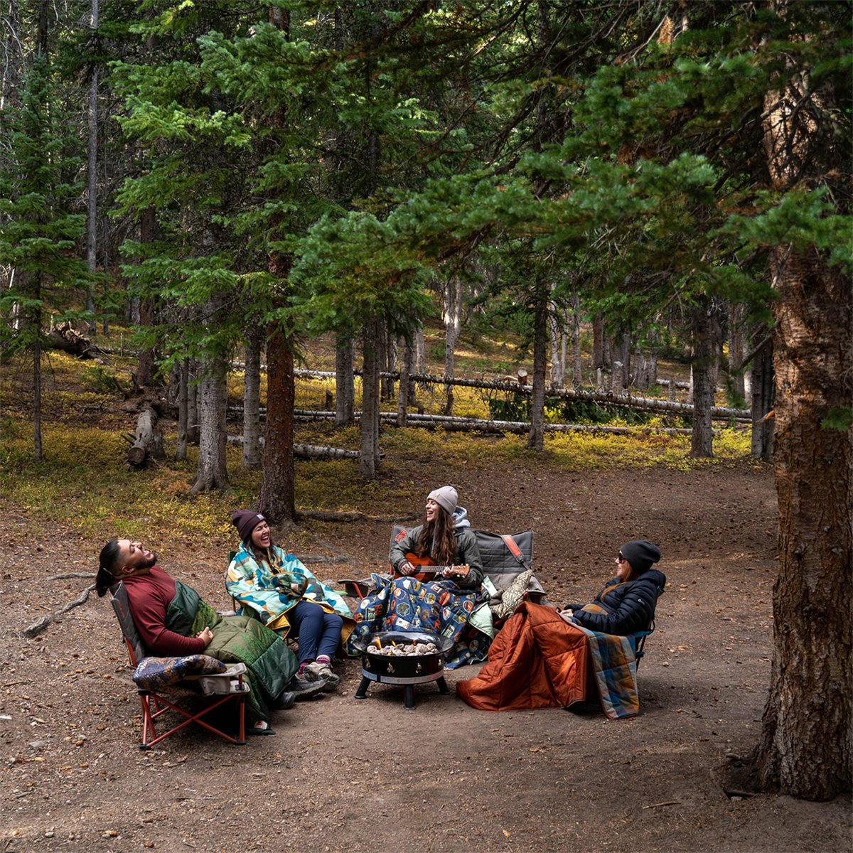 A group of people camp in the woods, laughing and sitting on the Kelty Loveseat and other Kelty chairs.