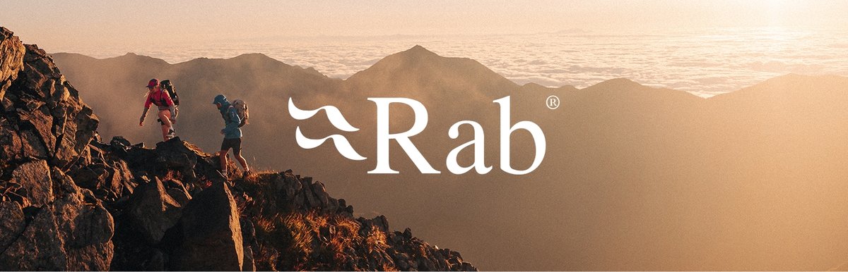 Two hikers in lightweight rain jackets traverse a rocky ridgeline during a hazy golden hour hike. The Rab logo appears in the middle of the horizon.