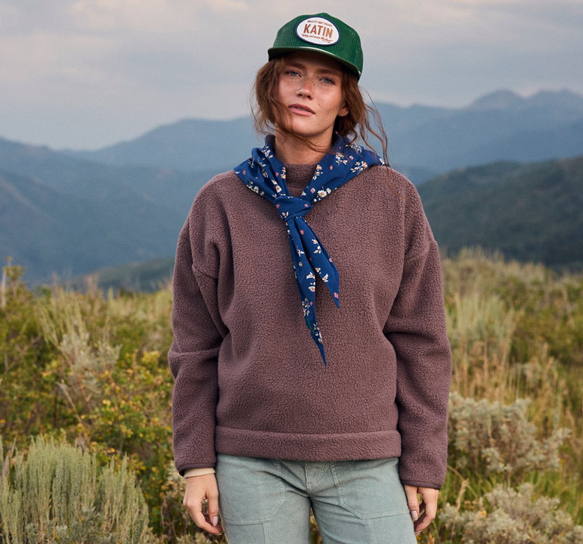 A woman stands against an alpine background, wearing the BGA Wander Mock Neck top.