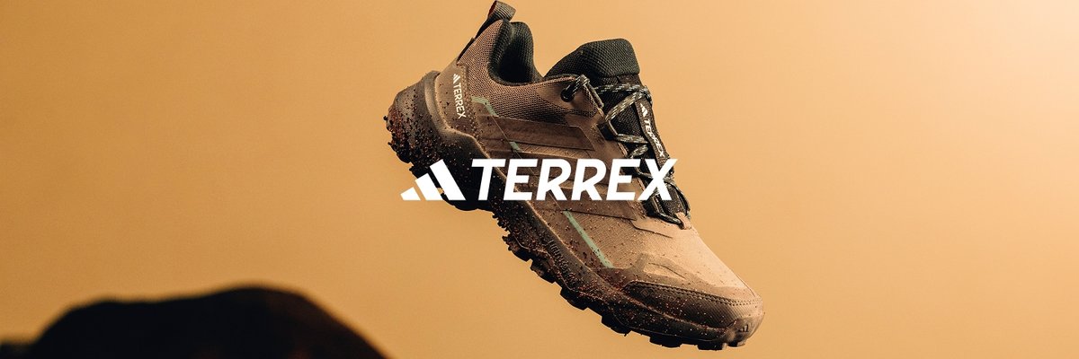 The adidas TERREX logo over a mud-splattered brown and black trail running shoe suspended in mid-air against a plain tan background.